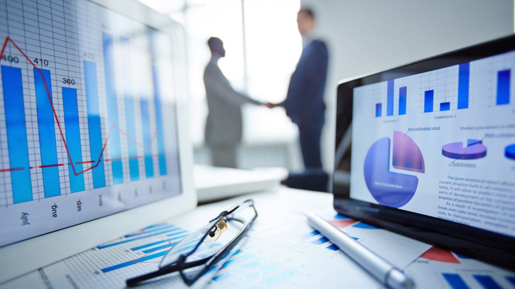 An image of eyeglasses, pen, two touchpads and financial documents at workplace with businessmen handshaking on background.